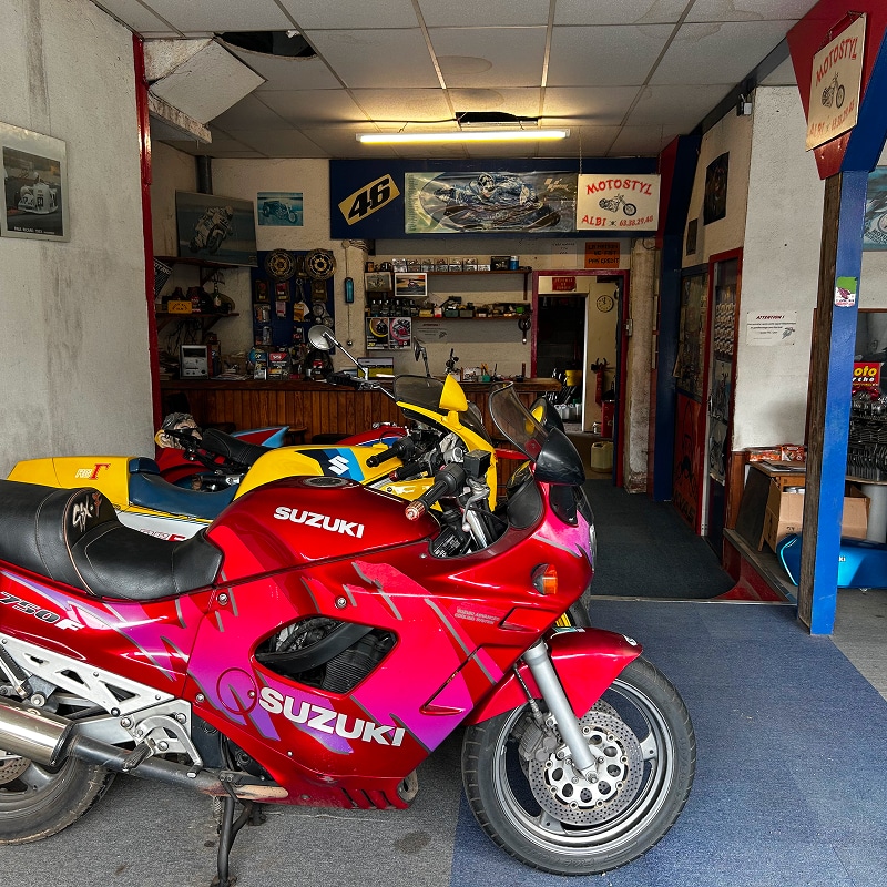 Moto Suzuki GSX-750F rouge et rose dans un garage vintage avec accessoires et affiches de course.