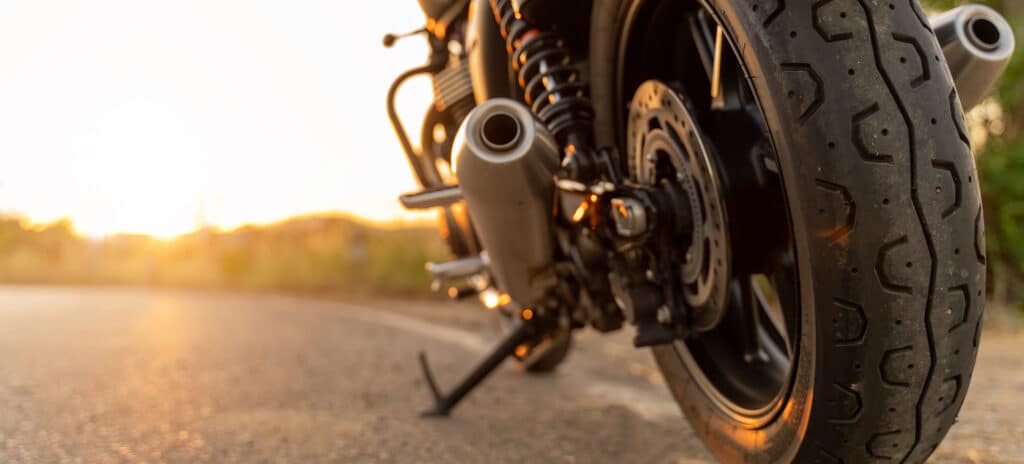 Gros plan sur le pneu et l'échappement d'une moto au coucher du soleil sur une route sinueuse.