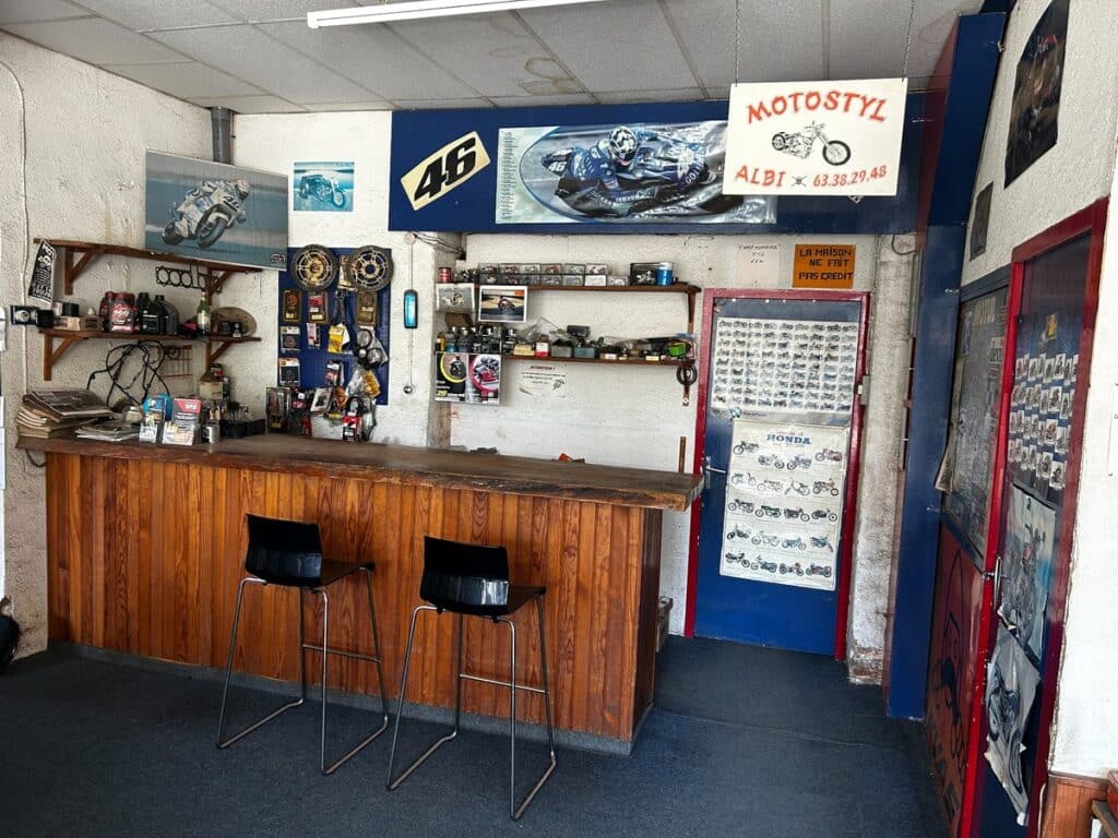 Garage Moto Motostyl Albi : Atelier et Pièces Racing Comptoir bois d'un atelier moto Motostyl à Albi avec affiches Valentino Rossi 46 et étagères de pièces mécaniques.